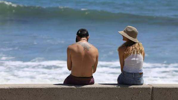 A couple sitting on a concrete wall looking out at the ocean on a sunny day