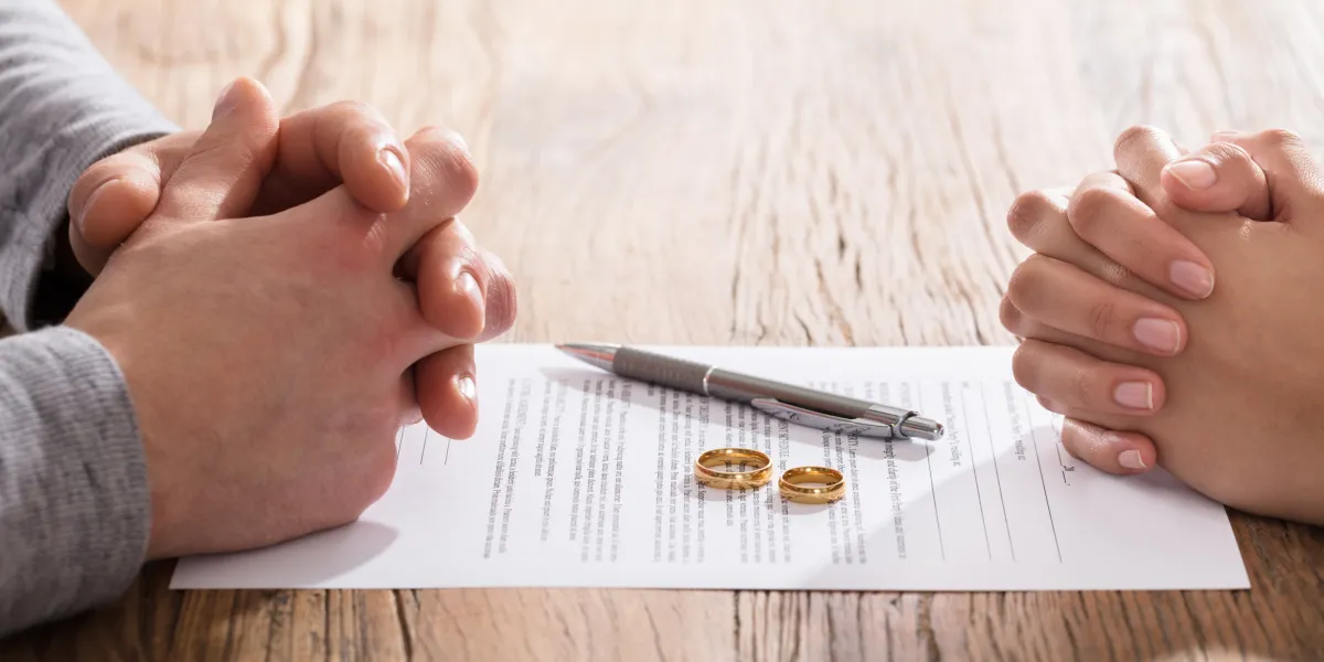 Two people with clasped hands across a table with divorce papers, pen and wedding rings