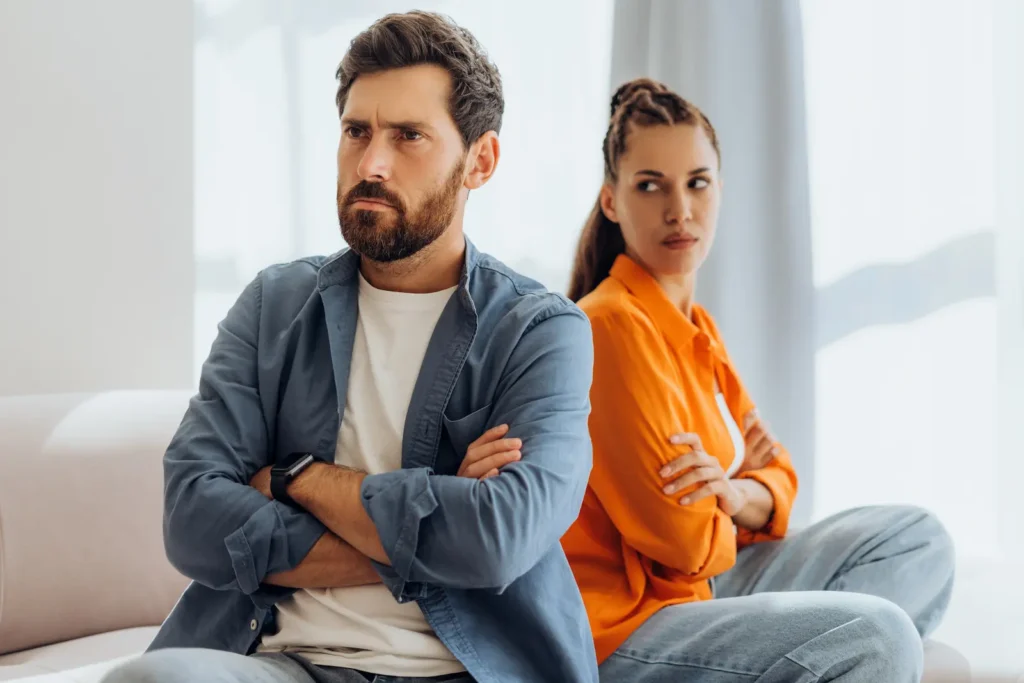 Couple sitting back-to-back on a couch with arms crossed, avoiding eye contact